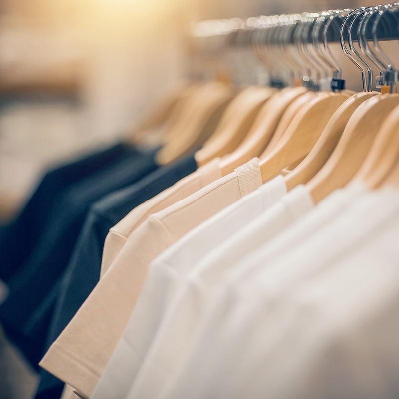 T-shirts unis beiges et blancs suspendus sur des cintres en bois sur une tringle métallique, éclairage doux et chaud.