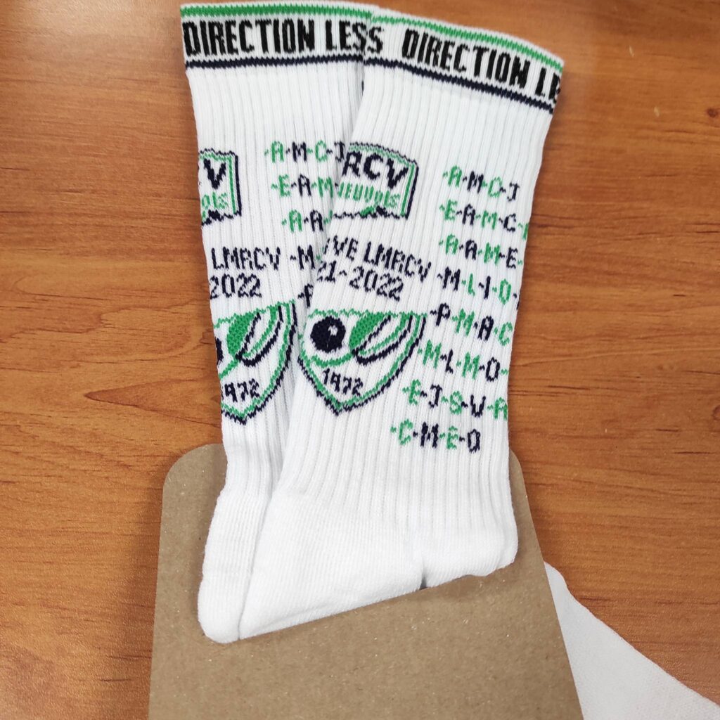 Paire de chaussettes blanches côtelées "DIRECTION LESS" avec logo sportif vert et noir centré surmontant l'année "1872". Acronymes et date "LMRCY 2022" brodés.