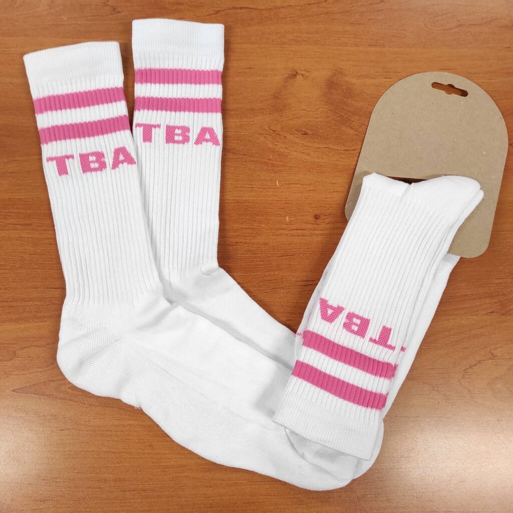 Trois chaussettes blanches montantes sport avec rayures roses et le texte "TBA". Elles sont présentées sur bois, dont une sur emballage carton.
