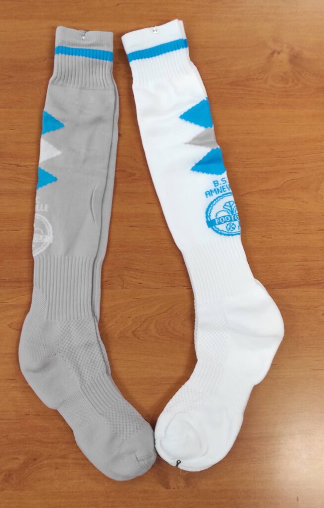 Chaussettes de football, kit domicile et extérieur (blanche et grise), posées sur bois. Motifs à chevrons bleus. Logo B.S. AMNEV FOOTBALL visible.