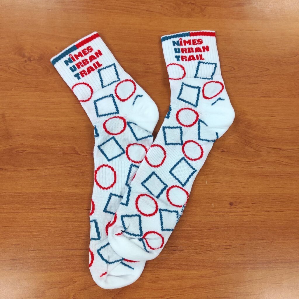 Paire de chaussettes blanches NÎMES URBAN TRAIL. Motifs graphiques géométriques bleus et rouges (carrés et cercles) sur fond bois.