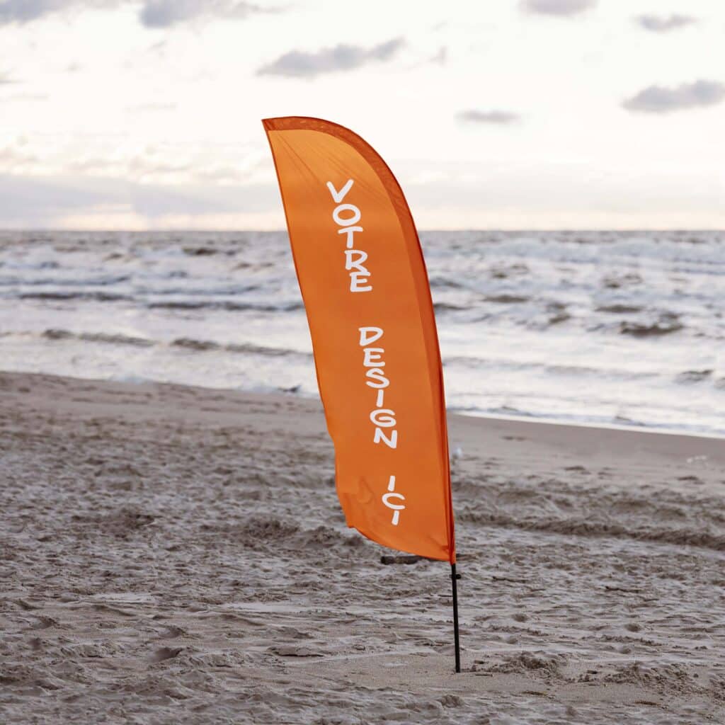 Drapeau Plume Orange sur la Plage : Affichez Votre Design Grand drapeau plume orange planté dans le sable d'une plage avec l'océan en arrière-plan. Le texte blanc dit "VOTRE DESIGN ICI".