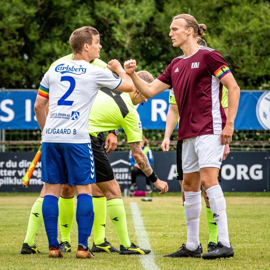 Joueurs de football : poignée de main et brassards Pride arc-en-ciel Deux joueurs de football se saluent (fist bump). Ils portent des brassards Pride arc-en-ciel. Maillots Carlsberg N°2 et bordeaux. VEJGAARD B visible.