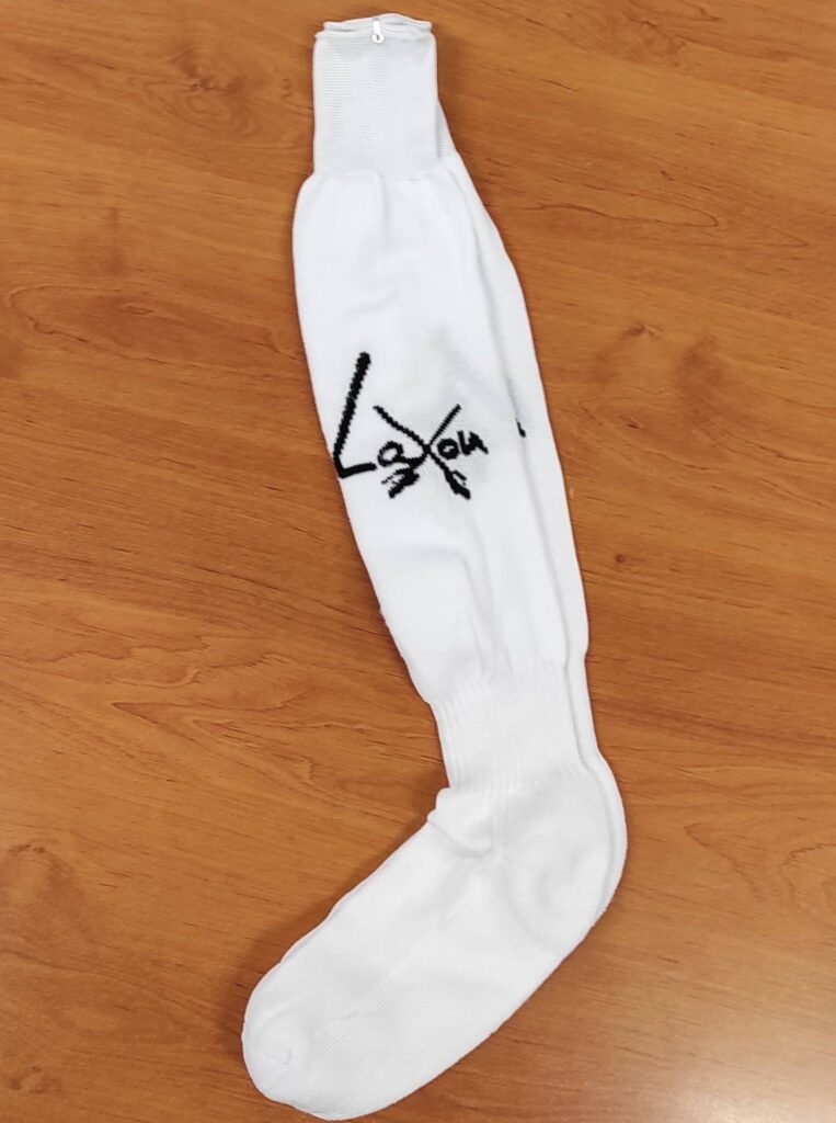 Chaussette longue de sport blanche avec le logo noir stylisé "Laxou" visible, posée sur un fond de bois brun clair.