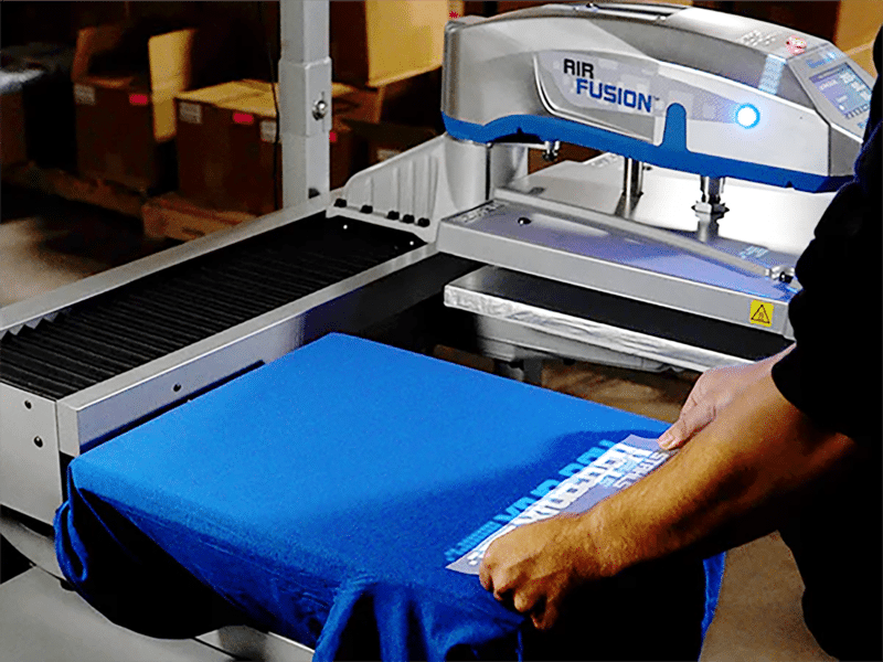 AIR FUSION : Presse à chaud professionnelle pour t-shirts. Application d'un transfert sur un t-shirt bleu. Presse à chaud automatique AIR FUSION en arrière-plan, prête pour le marquage textile.