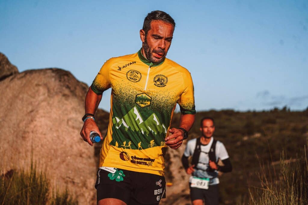 Trail running intense : coureur déterminé en montagne Trail runner concentré et en sueur, en maillot jaune/vert (JUVA, ADFA), bouteille à la main, courant sur un sentier rocheux.