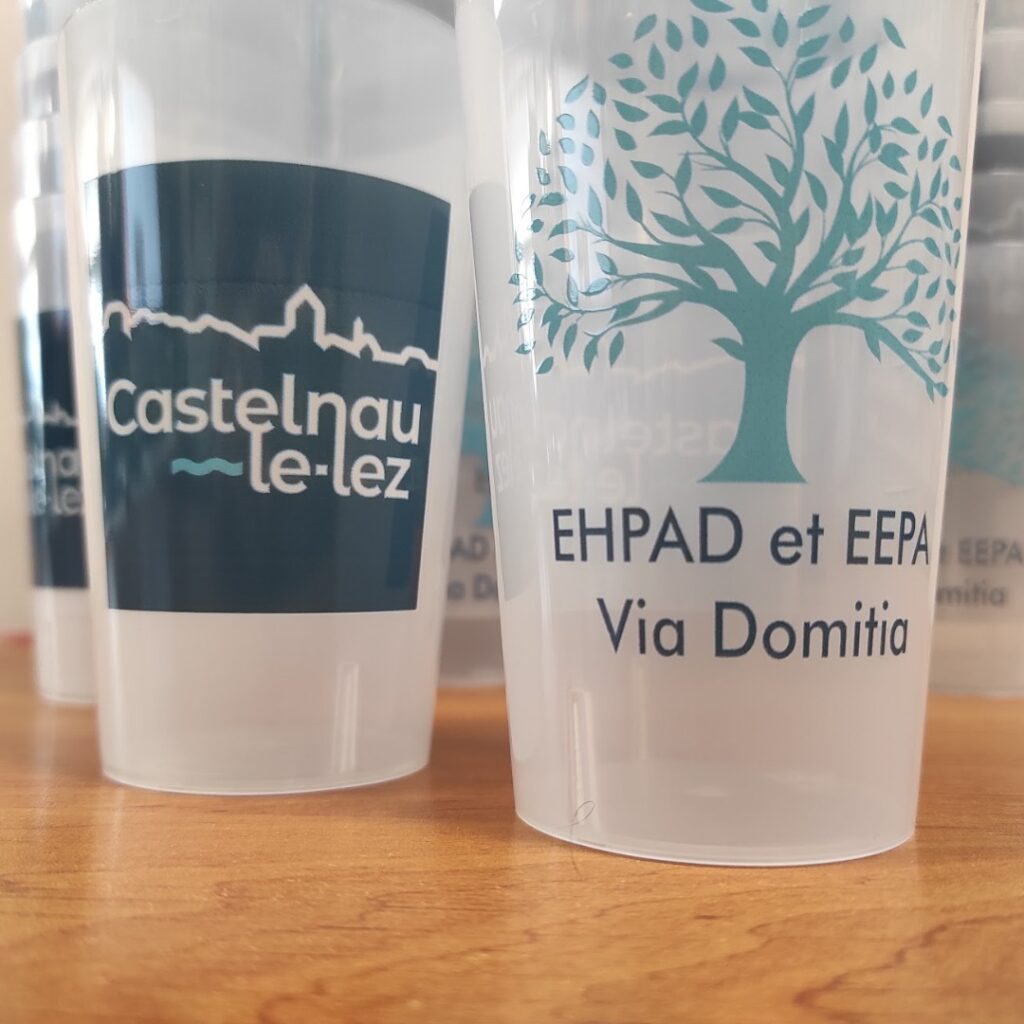 Gobelets réutilisables avec logos Castelnau-le-Lez et EHPAD Via Domitia posés sur une table en bois.
